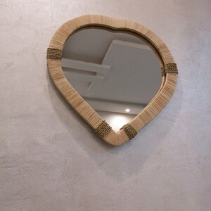 May include: A heart-shaped mirror with a woven frame made of natural materials. The frame is accented with a brown rope detail.