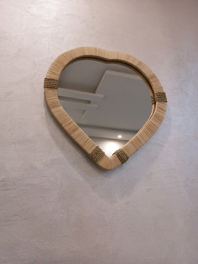 May include: A heart-shaped mirror with a woven frame made of natural materials. The frame is accented with a brown rope detail.