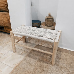 May include: A white rope bench with a natural wood frame. The bench is sitting on a light brown rug in a room with white walls.