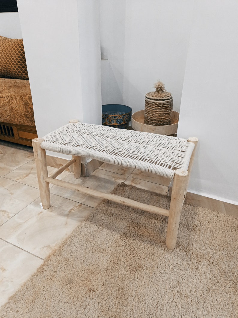 May include: A white rope bench with a natural wood frame. The bench is sitting on a light brown rug in a room with white walls.