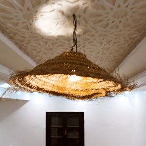 May include: A woven, straw lampshade hangs from a chain, illuminating a room with a white ceiling and white walls. The lampshade is round and has a natural, light brown color.