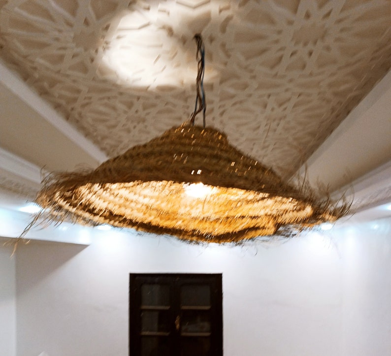 May include: A woven, straw lampshade hangs from a chain, illuminating a room with a white ceiling and white walls. The lampshade is round and has a natural, light brown color.