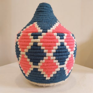 Könnte beinhalten: Ein geflochtener Korb mit einem geometrischen Muster in Blau, Rosa und Weiß. Der Korb ist rund und hat eine große Öffnung.