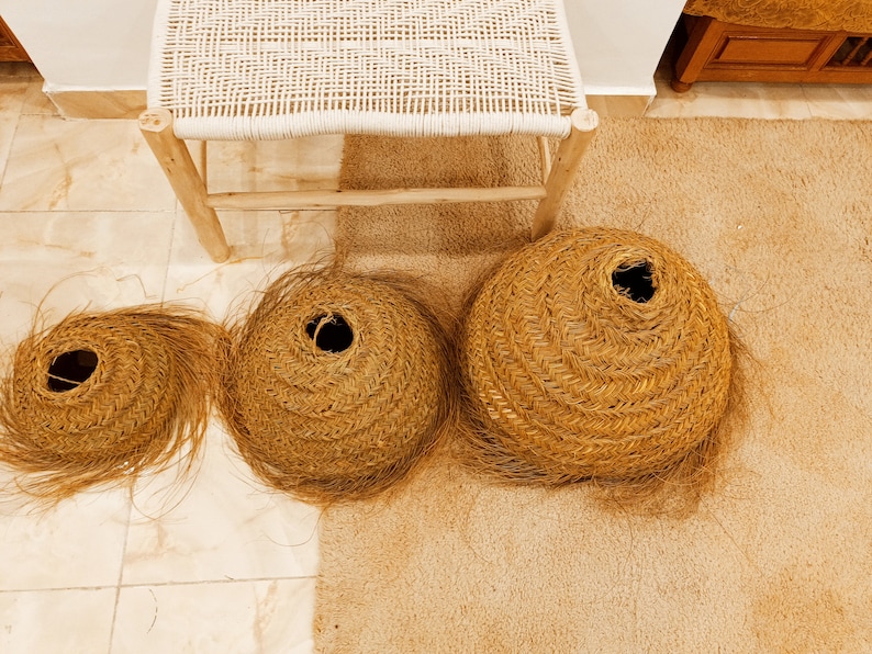 K&ouml;nnte beinhalten: Drei gewebte Strohk&ouml;rbe in verschiedenen Gr&ouml;&szlig;en, jeweils mit einer zentralen &Ouml;ffnung. Die K&ouml;rbe sind warmbraun und haben ein Spiraldesign. Im Hintergrund steht ein kleiner Holzstuhl mit gewebter Sitzfl&auml;che.