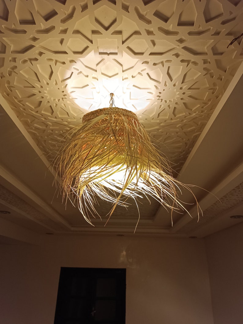 K&ouml;nnte beinhalten: Eine geflochtene, stroh&auml;hnliche Pendelleuchte h&auml;ngt an einer Decke mit einem dekorativen, wei&szlig;en geometrischen Muster. Die Leuchte ist beleuchtet und strahlt ein warmes Licht aus.