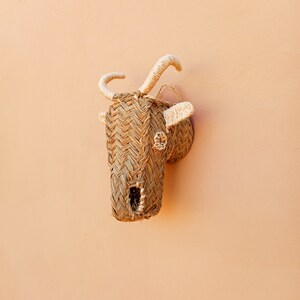 May include: A woven straw animal head wall decor in the shape of a goat with white rope horns. The goat head is mounted on a light peach wall.