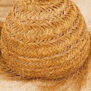 K&ouml;nnte beinhalten: Ein geflochtener Korb mit rundem Boden und konischem Oberteil. Der Korb besteht aus naturfarbenem Stroh oder getrocknetem Gras mit einem eng gewebten Muster. Die Textur ist sichtbar, und einige lose Str&auml;hnen erstrecken sich von der Basis.