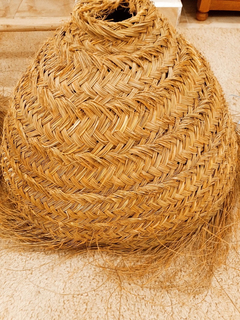 K&ouml;nnte beinhalten: Ein geflochtener Korb mit rundem Boden und konischem Oberteil. Der Korb besteht aus naturfarbenem Stroh oder getrocknetem Gras mit einem eng gewebten Muster. Die Textur ist sichtbar, und einige lose Str&auml;hnen erstrecken sich von der Basis.