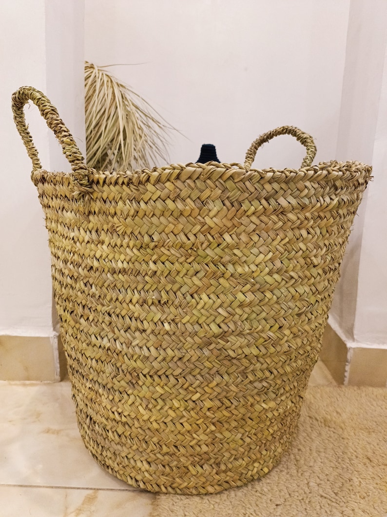 May include: A large, natural woven basket with two handles. The basket is made of a light brown, natural material and has a woven pattern.
