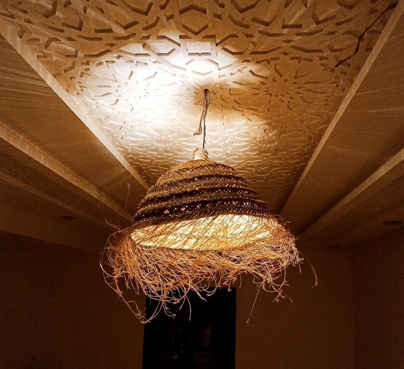 K&ouml;nnte beinhalten: Eine H&auml;ngeleuchte mit einem gewebten, braunen Lampenschirm. Das Licht ist eingeschaltet und erzeugt einen warmen Schein. Die Decke dar&uuml;ber weist ein strukturiertes, geometrisches Muster auf. Die Leuchte hat ein nat&uuml;rliches, organisches Design.