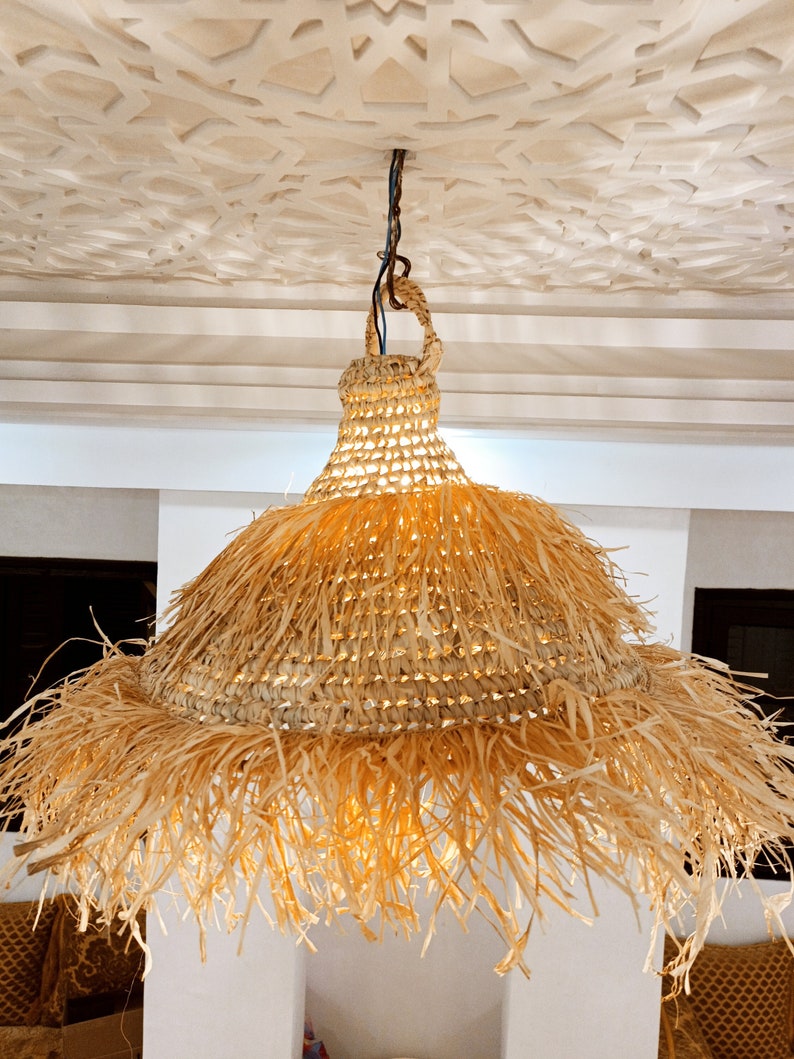 May include: A large, round, woven pendant light fixture with a natural straw color and a fringe border. The light fixture is hanging from a ceiling with a decorative pattern.