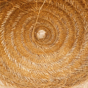 K&ouml;nnte beinhalten: Nahaufnahme eines geflochtenen Strohhuts, der ein Spiralmuster zeigt. Der Hut besteht aus nat&uuml;rlichem, hellbraunem Stroh mit einem zentralen Loch. Das Geflecht erzeugt eine strukturierte Oberfl&auml;che, und der Hut liegt auf einer hellen Oberfl&auml;che.