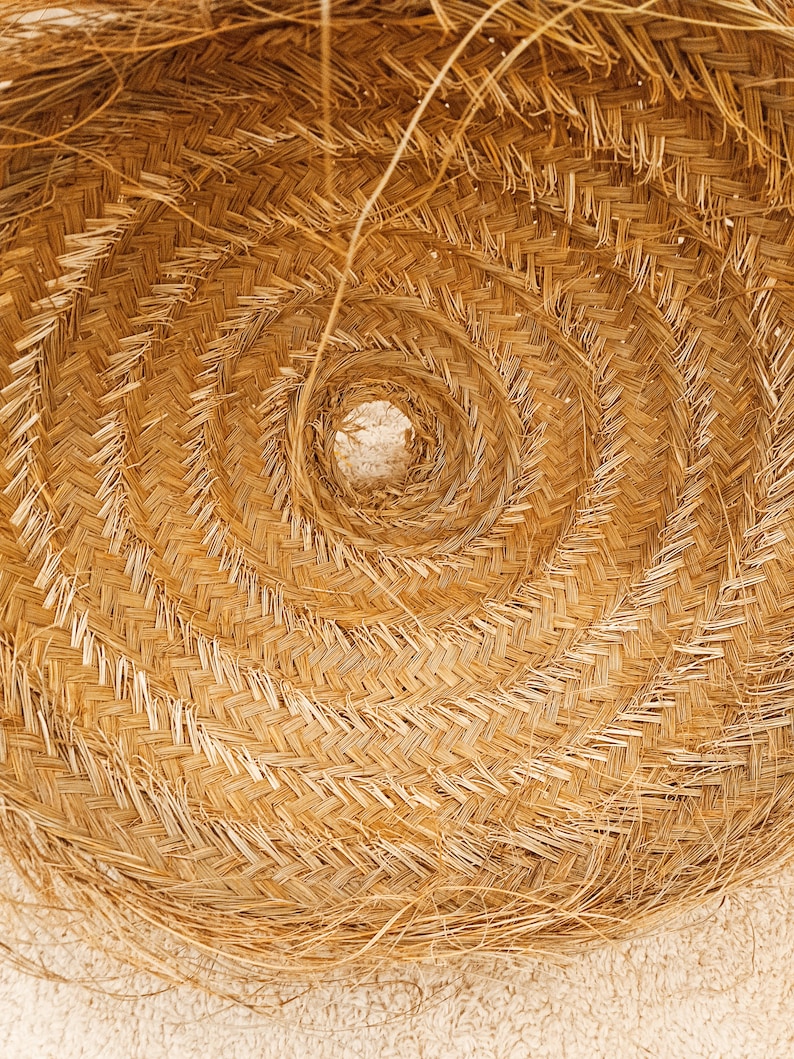K&ouml;nnte beinhalten: Nahaufnahme eines geflochtenen Strohhuts, der ein Spiralmuster zeigt. Der Hut besteht aus nat&uuml;rlichem, hellbraunem Stroh mit einem zentralen Loch. Das Geflecht erzeugt eine strukturierte Oberfl&auml;che, und der Hut liegt auf einer hellen Oberfl&auml;che.