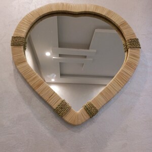 May include: A heart-shaped mirror with a woven rattan frame and green accents. The mirror is hanging on a white wall.