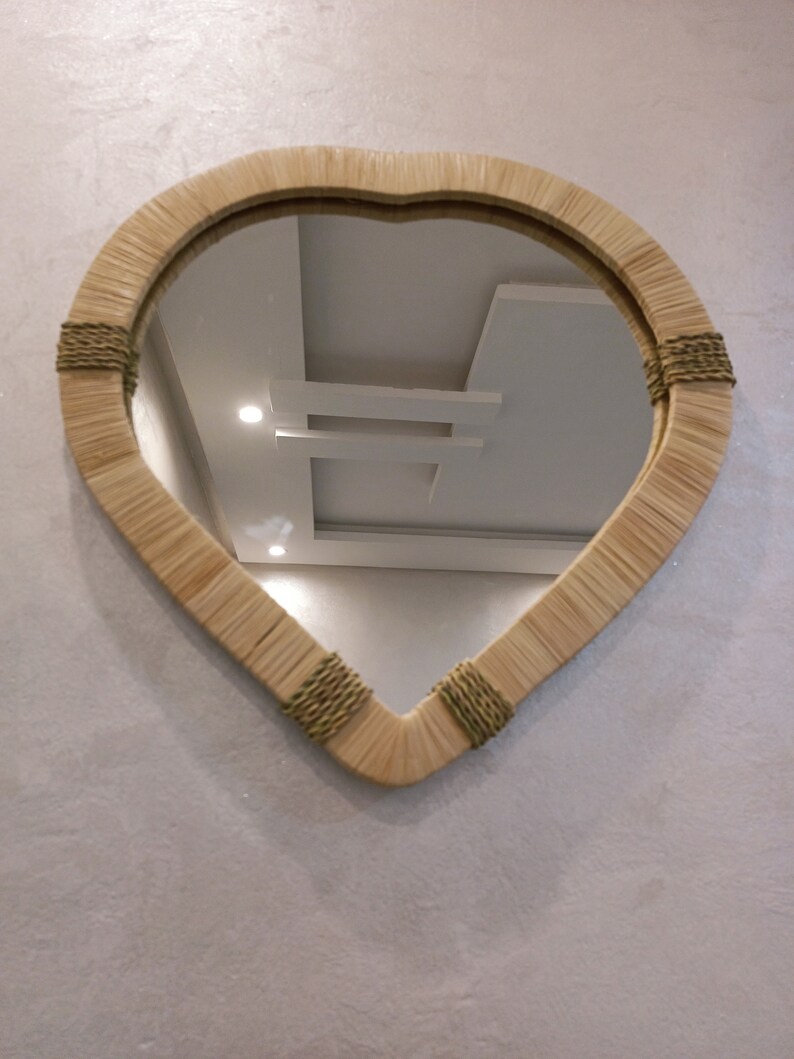 May include: A heart-shaped mirror with a woven rattan frame and green accents. The mirror is hanging on a white wall.