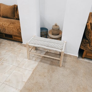 May include: A white woven bench with wooden legs sits on a beige rug in a room with white walls. A small blue and white patterned stool and a woven basket with a lid are visible in the background.