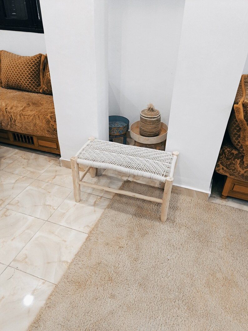 May include: A white woven bench with wooden legs sits on a beige rug in a room with white walls. A small blue and white patterned stool and a woven basket with a lid are visible in the background.