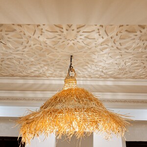 May include: A large, round, woven, straw lampshade hangs from a ceiling with a white, intricate, geometric pattern. The lampshade is made of natural, light brown, woven straw with a fringe around the bottom.