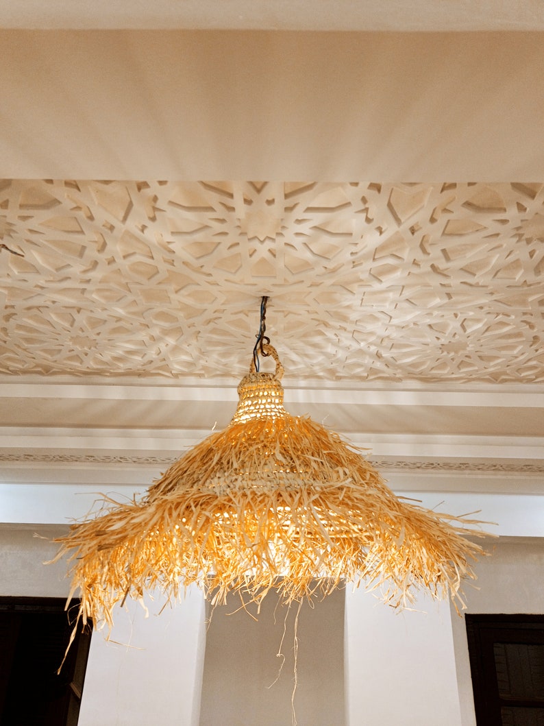 May include: A large, round, woven, straw lampshade hangs from a ceiling with a white, intricate, geometric pattern. The lampshade is made of natural, light brown, woven straw with a fringe around the bottom.