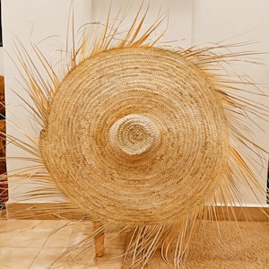 May include: A large, round, woven straw hat with a wide brim and a natural brown colour. The hat is sitting on a wooden stool.