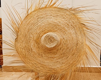 May include: A large, round, woven straw hat with a wide brim and a natural brown color. The hat is sitting on a wooden stool.