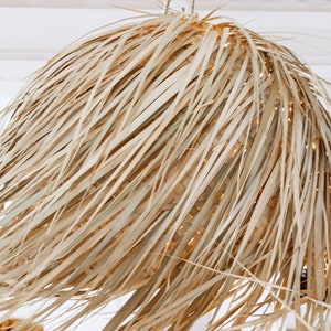 May include: A large, round, woven pendant light made of natural, light brown, dried palm leaves. The light is hanging from a white ceiling.