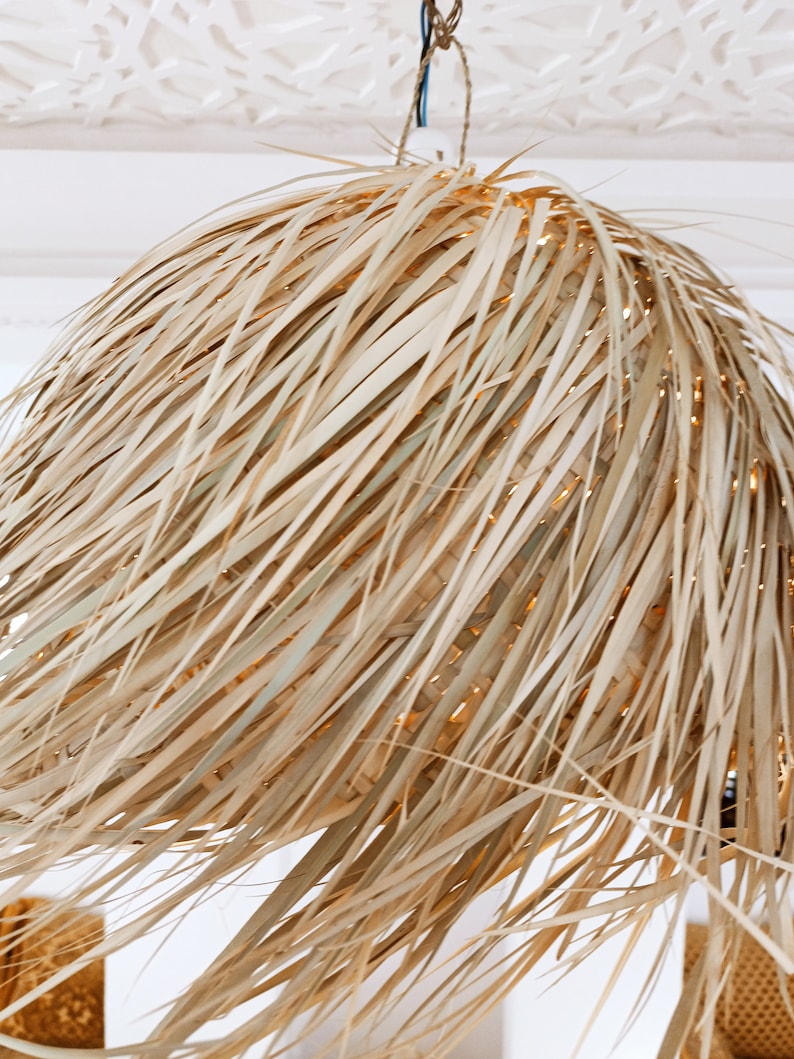 May include: A large, round, woven pendant light made of natural, light brown, dried palm leaves. The light is hanging from a white ceiling.
