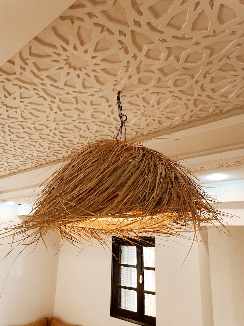 May include: A large, round, woven light fixture hangs from a ceiling with a decorative, white, geometric pattern. The fixture is made of natural materials and has a light brown color.
