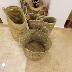 May include: Three woven baskets made of natural materials. The baskets are different sizes and shapes, with two round baskets and one tall, cylindrical basket. The baskets are sitting on a light brown rug.