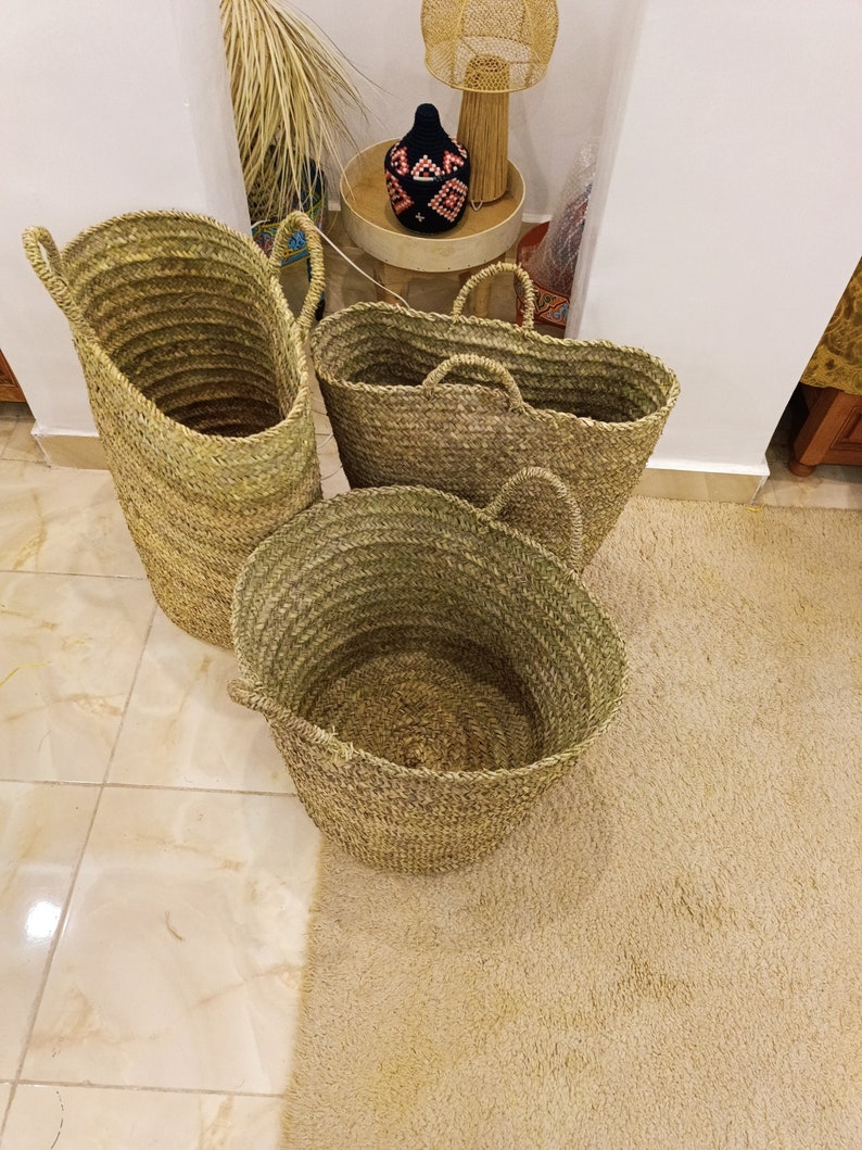 May include: Three woven baskets made of natural materials. The baskets are different sizes and shapes, with two round baskets and one tall, cylindrical basket. The baskets are sitting on a light brown rug.