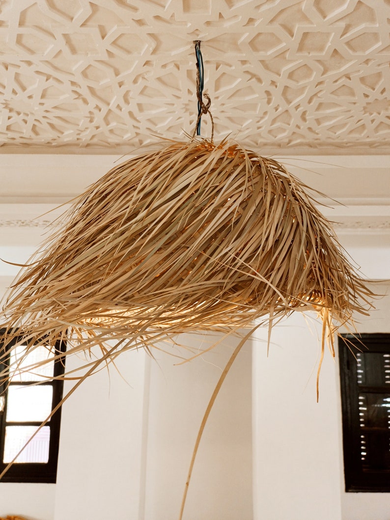 May include: A large, round, natural woven pendant light fixture hangs from a white ceiling with a decorative pattern. The light fixture is made of woven natural fibers, giving it a rustic and bohemian aesthetic.