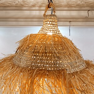 May include: A woven straw pendant light with a fringed bottom. The light is hanging from a black cord and is lit.