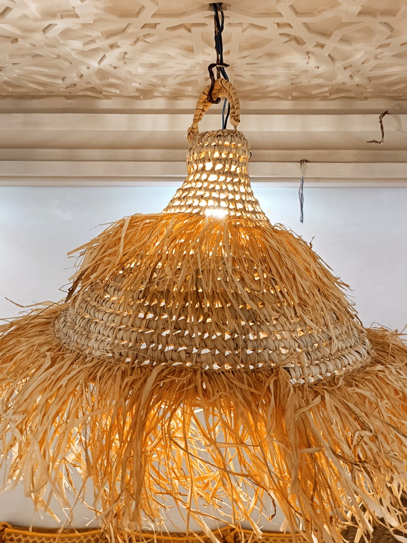 May include: A woven straw pendant light with a fringed bottom. The light is hanging from a black cord and is lit.