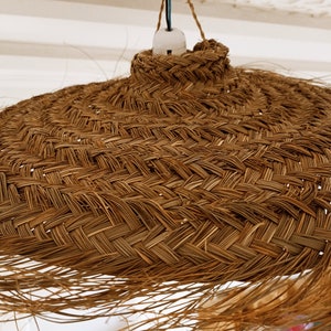 Może przedstawiać: Pleciona lampa wisząca z wikliny w naturalnym brązowym kolorze. Lampa ma szeroki, okrągły kształt i jest wykonana z naturalnych materiał&oacute;w.