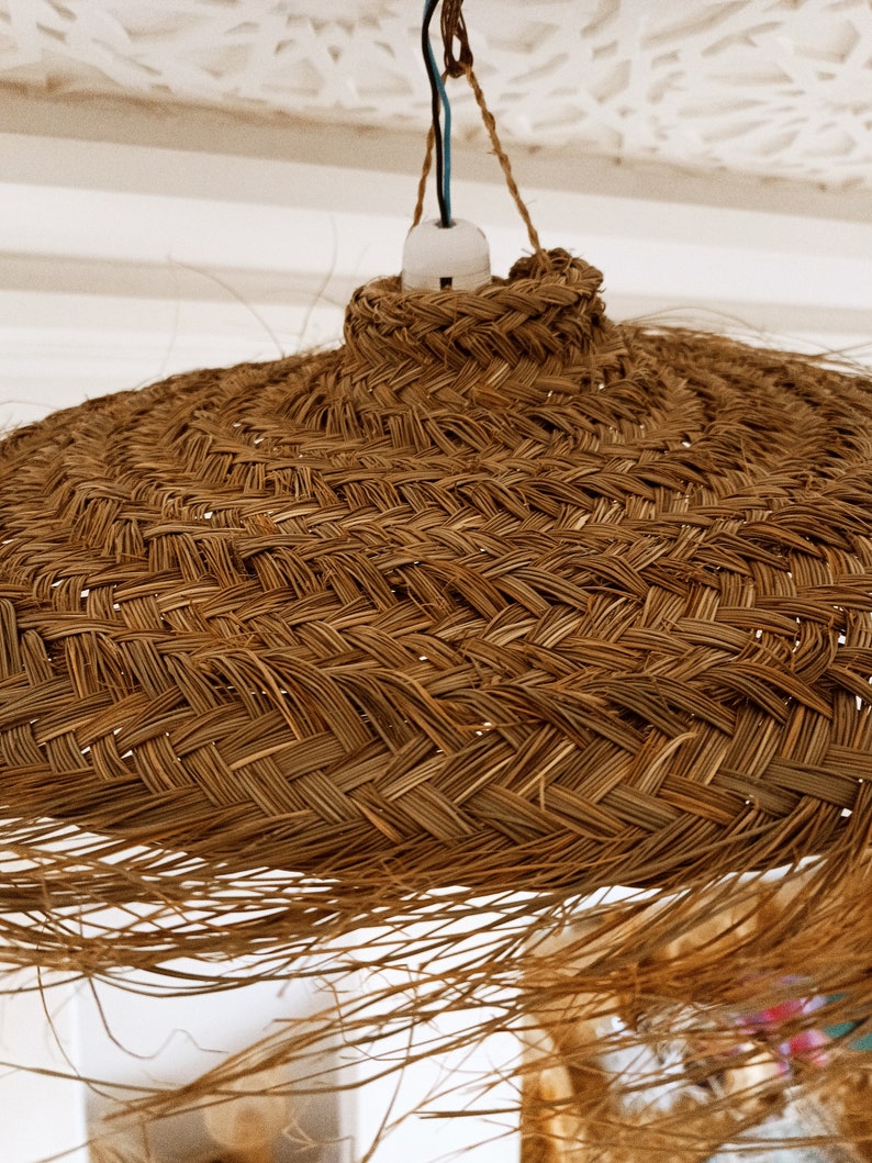 Może przedstawiać: Pleciona lampa wisząca z wikliny w naturalnym brązowym kolorze. Lampa ma szeroki, okrągły kształt i jest wykonana z naturalnych materiał&oacute;w.
