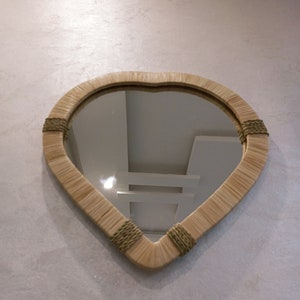 May include: A heart-shaped mirror with a woven frame in a light brown color. The frame has green accents. The mirror is reflecting a white ceiling with light fixtures.