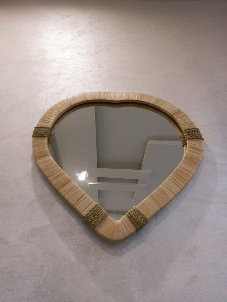 May include: A heart-shaped mirror with a woven frame in a light brown color. The frame has green accents. The mirror is reflecting a white ceiling with light fixtures.