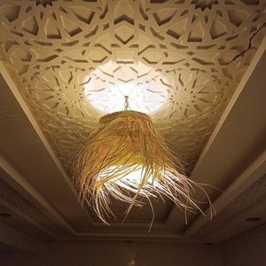 K&ouml;nnte beinhalten: Eine geflochtene, stroh&auml;hnliche Pendelleuchte h&auml;ngt an einer Decke mit einem dekorativen, wei&szlig;en geometrischen Muster. Die Leuchte strahlt ein warmes Licht aus.