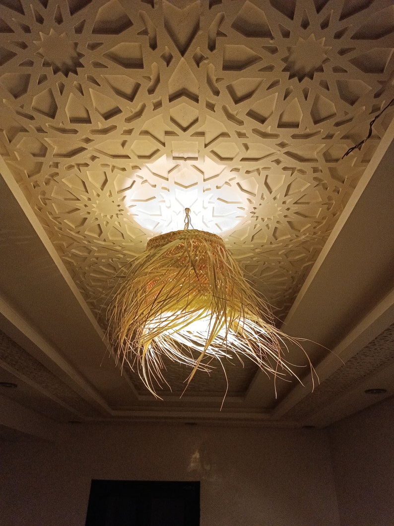 K&ouml;nnte beinhalten: Eine geflochtene, stroh&auml;hnliche Pendelleuchte h&auml;ngt an einer Decke mit einem dekorativen, wei&szlig;en geometrischen Muster. Die Leuchte strahlt ein warmes Licht aus.