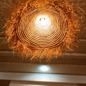 May include: A round, woven, straw lampshade with a light bulb inside. The lampshade is hanging from the ceiling and has a fringe of straw around the bottom.