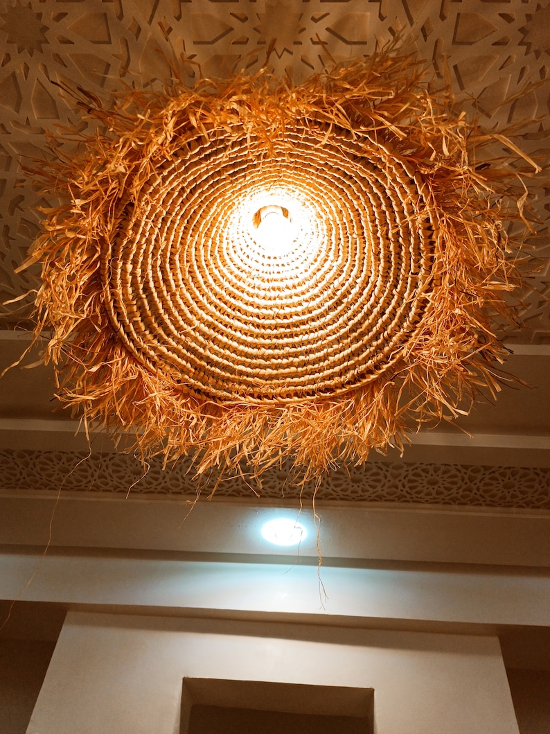 May include: A round, woven, straw lampshade with a light bulb inside. The lampshade is hanging from the ceiling and has a fringe of straw around the bottom.