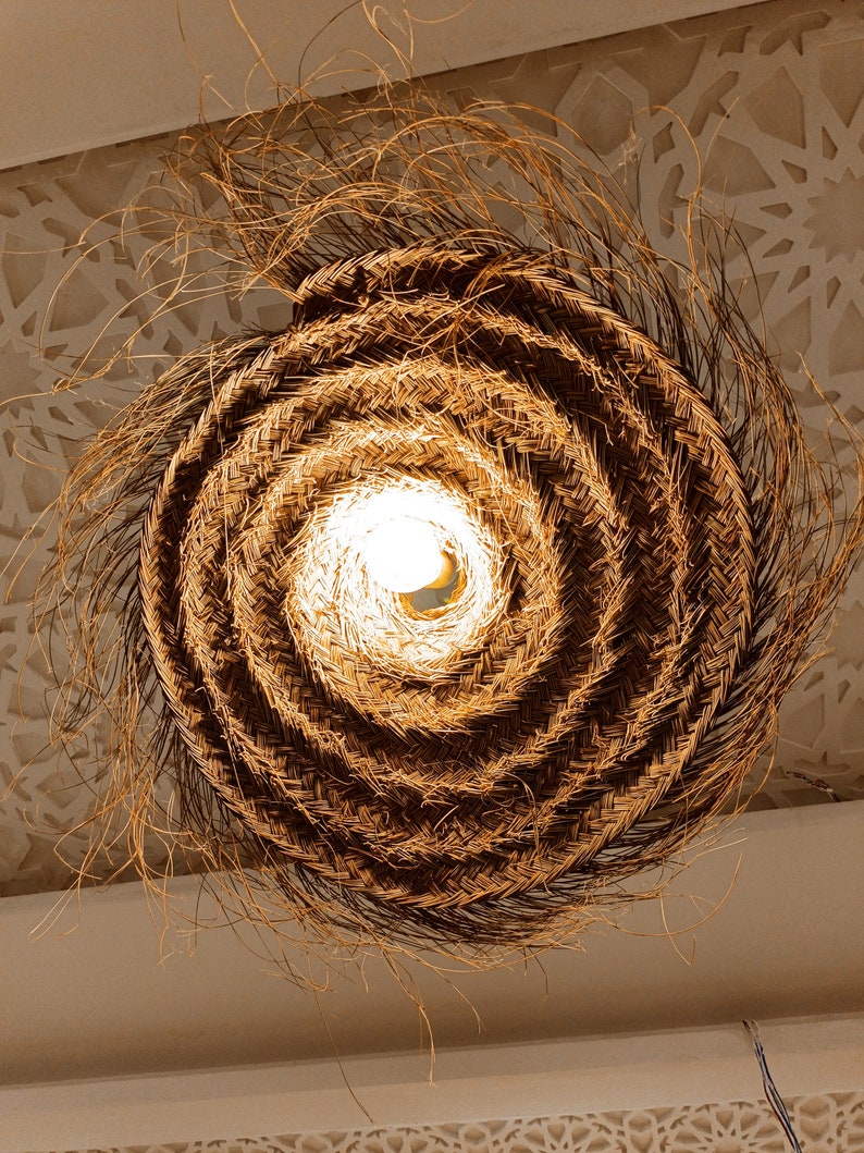 Może przedstawiać: Okrągła lampa wykonana z plecionki w naturalnym brązowym kolorze. Lampa jest wykonana z cienkich, splecionych pasm materiału i ma żar&oacute;wkę w środku.