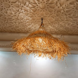 May include: A large, round, straw lampshade hangs from a ceiling with a decorative, white, patterned design. The lampshade is made of woven straw with a fringe around the bottom.