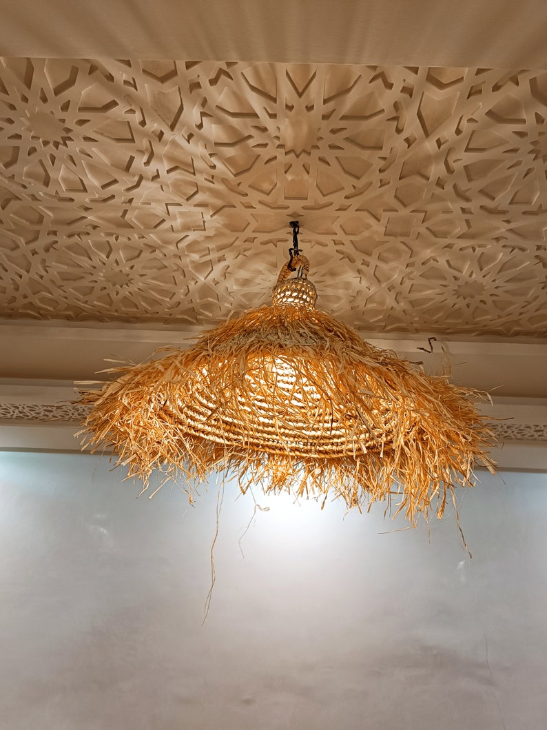 May include: A large, round, straw lampshade hangs from a ceiling with a decorative, white, patterned design. The lampshade is made of woven straw with a fringe around the bottom.