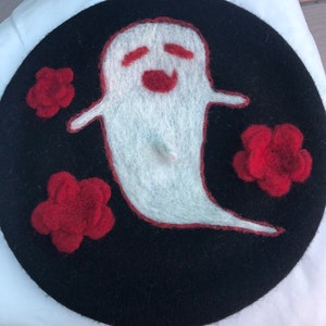 Puede incluir: Un gorro negro con un fantasma de fieltro blanco y tres flores de fieltro rojo. El fantasma tiene una cara sonriente.