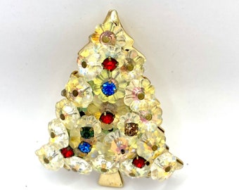 May include: A vintage Christmas tree brooch. The brooch is shaped like a Christmas tree and is covered in clear, iridescent rhinestones. The tree is decorated with red, blue, and brown rhinestones. The brooch has a gold-tone metal backing.