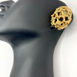 Puede incluir: Pendiente dorado de clip con diseño tejido. El pendiente es redondo y tiene una apariencia tridimensional. Se muestra en una cabeza de maniquí negra sobre un fondo blanco. Accesorio vintage.