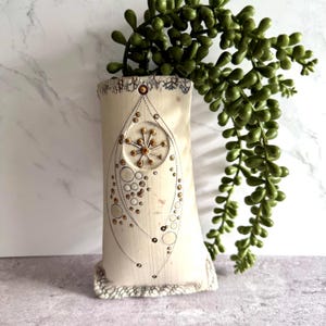 Può includere: Vaso in ceramica color crema con un design decorativo di cerchi e punti marroni e dorati. Il vaso ha un bordo e una base strutturati. Steli di piante verdi e frondosi cadono dalla parte superiore del vaso.