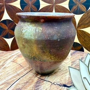 May include: A rustic, handcrafted earthenware pot with a wide mouth and rounded body. The pot features a blend of earthy tones, including brown, green, and reddish-brown hues. The exterior has a textured, aged appearance, suggesting it's an antique or vintage piece.