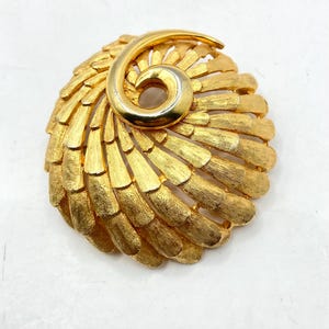 Puede incluir: Broche vintage dorado con un diseño texturizado en forma de abanico. El broche presenta un acento en espiral en la parte superior, agregando un toque único. Una pieza llamativa.